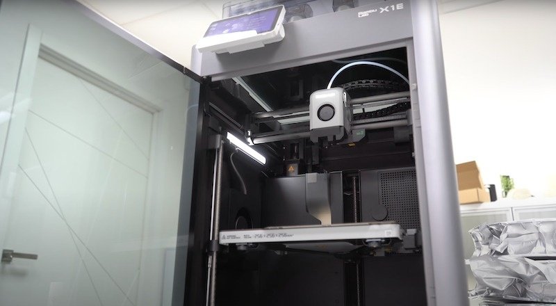 The Bambu X1E 3D printer is showcased with its build chamber door open, revealing the internal components. The printer features a sleek, vertical design with a metallic frame. A mounted display screen is at the top, angled for user interaction. Inside, the extruder and filament system are visible, with a spool holder and filament guide tube. The printer is situated in a modern room setting with a glass door in the background.