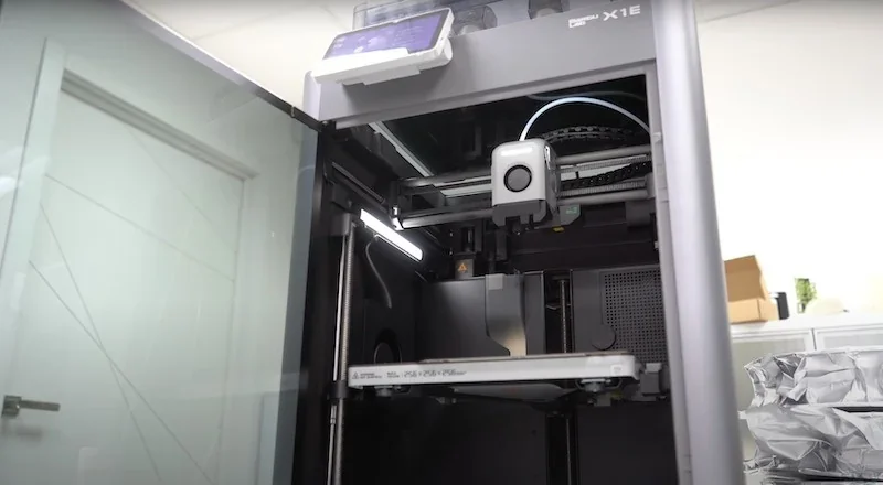 Bambu X1E 3D printer in an office, with open door showing internal components and printed samples on a shelf.