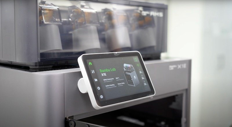This image presents an external view of the Bambu X1E 3D printer with a focus on its user interface. The printer has a touchscreen control panel displaying operational information such as 