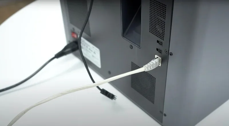 Bambu X1E rear view showing Ethernet cable connected, power cord plugged in, on a white surface, black-grey casing with ventilation panels visible.