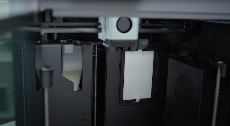The interior view of the Bambu X1E 3D printer featuring its mechanical and electronic components. The printer's interior showcases dual lead screws on either side for stable movement. The central section contains a print head assembly with a precision nozzle system. Visible is a filtration component encased in a frame, suggesting advanced air management for clean prints. The design emphasizes precision engineering with a focus on maintaining a controlled printing environment.