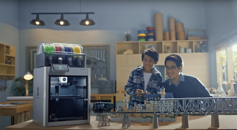The Bambu X1E 3D printer is prominently featured in a home workshop setting, positioned on a wooden table. The printer's multi-color filament storage is visible on top, underscoring its multi-material printing capability. In the background, a child and an adult share enthusiasm over a detailed miniature train set and architectural diorama, exemplifying 3D-printed creations by the Bambu X1E. The well-lit room with shelves and decorative items creates a warm, inviting atmosphere for creativity.