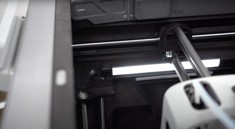 The image showcases the internal mechanics of the Bambu X1E 3D printer, focusing on the precision components within the machine. Visible is the gantry system, responsible for guiding the print head, along with internal lighting that enhances visibility. There are control systems labeled with "OPEN," indicating accessibility features for maintenance or adjustments. This detailed view highlights the robust engineering and design intended for efficient and accurate 3D printing.