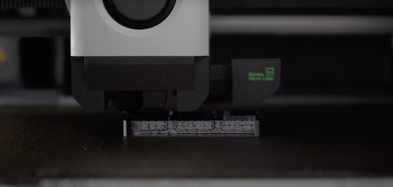 A close-up view of the Bambu X1E 3D printer's extruder in action. The image captures the extruder meticulously layering black filament onto the print bed, demonstrating the precision and accuracy of the printer. The branding on the printer is subtly visible, with a small green emblem reading 