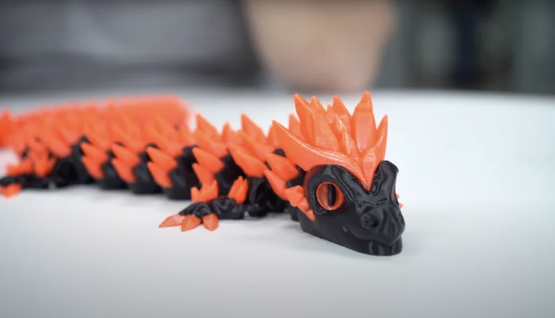 Orange and black 3D-printed dragon figurine with spiky texture, crafted by the Bambu X1E, displayed on a white surface.