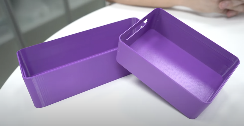 Two purple storage containers are displayed on a white surface, created by the Bambu Lab X1E 3D printer. They have a rectangular shape with smooth, glossy surfaces and slightly rounded edges. One container is inverted to show its spacious interior. The high-quality finish and consistent thickness demonstrate the printer's capability to produce precise and functional everyday objects.
