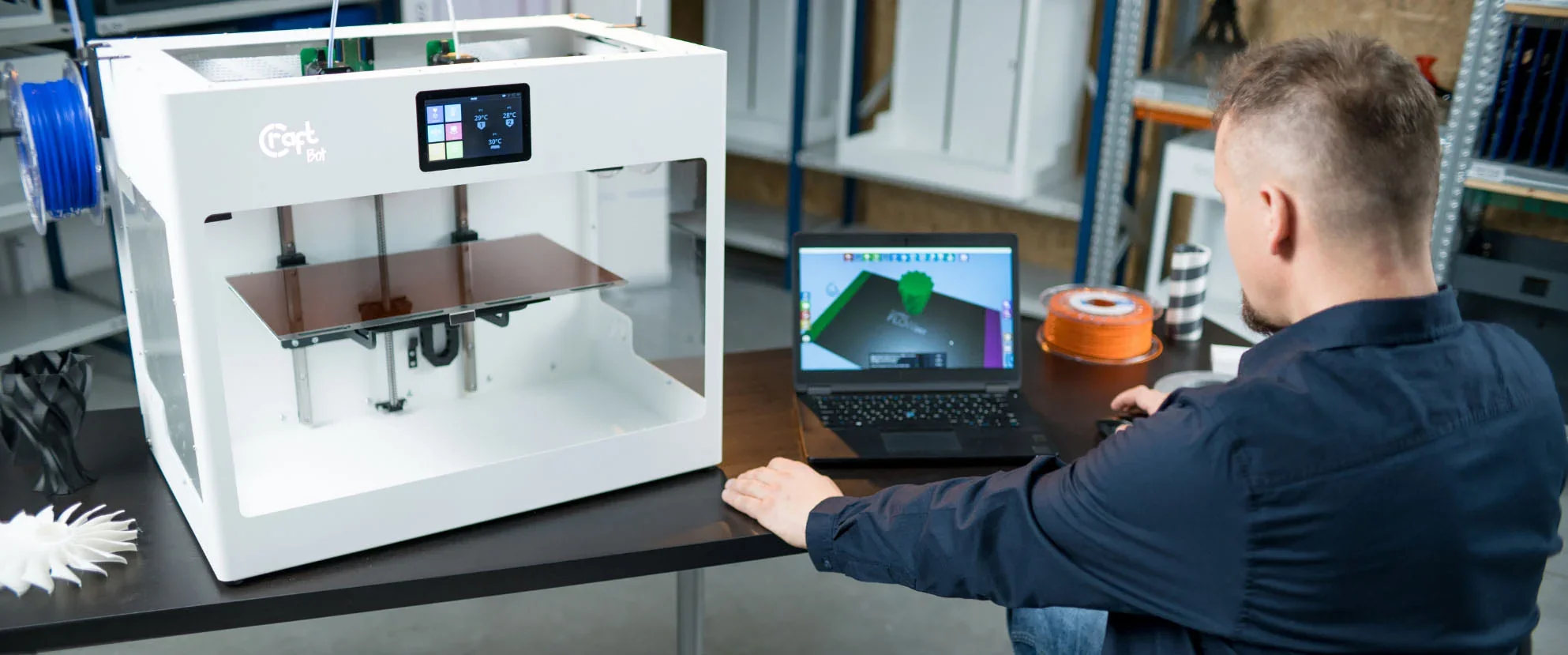 CraftBot Flow IDEX XL 3D printer on a desk, printing displayed on connected laptop, filament spool, created objects nearby, man operating setup.