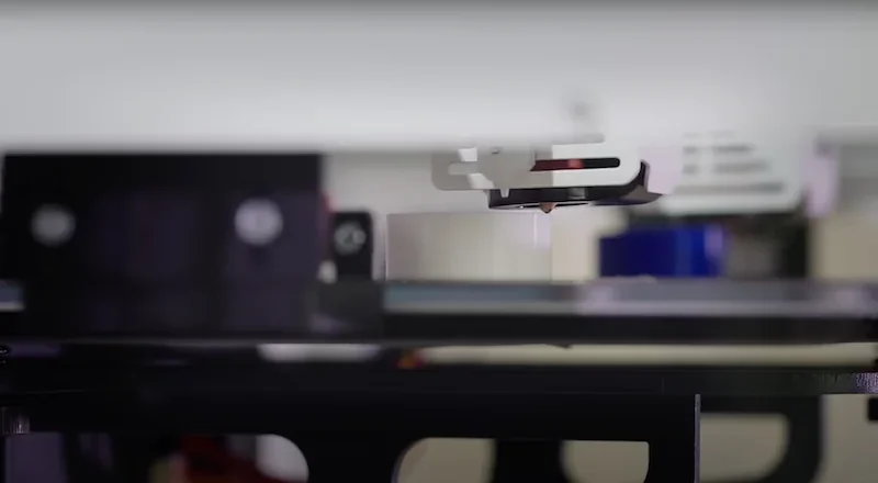 Craftbot Flow IDEX XL 3D printer close-up, depicts nozzle printing white object on glass bed, machine in operation, intricate detail visible.