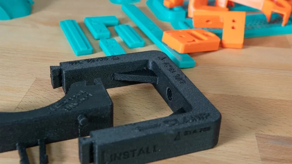 3D-printed parts in black, teal, and orange on a wooden surface, showcasing Creality K1's capabilities with varied shapes and colors.