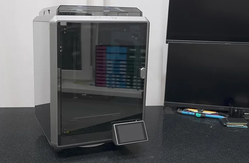Creality K1 3D printer on a desk with a control screen, sleek design, accompanied by pens and monitor, showcasing its modern, efficient setup.