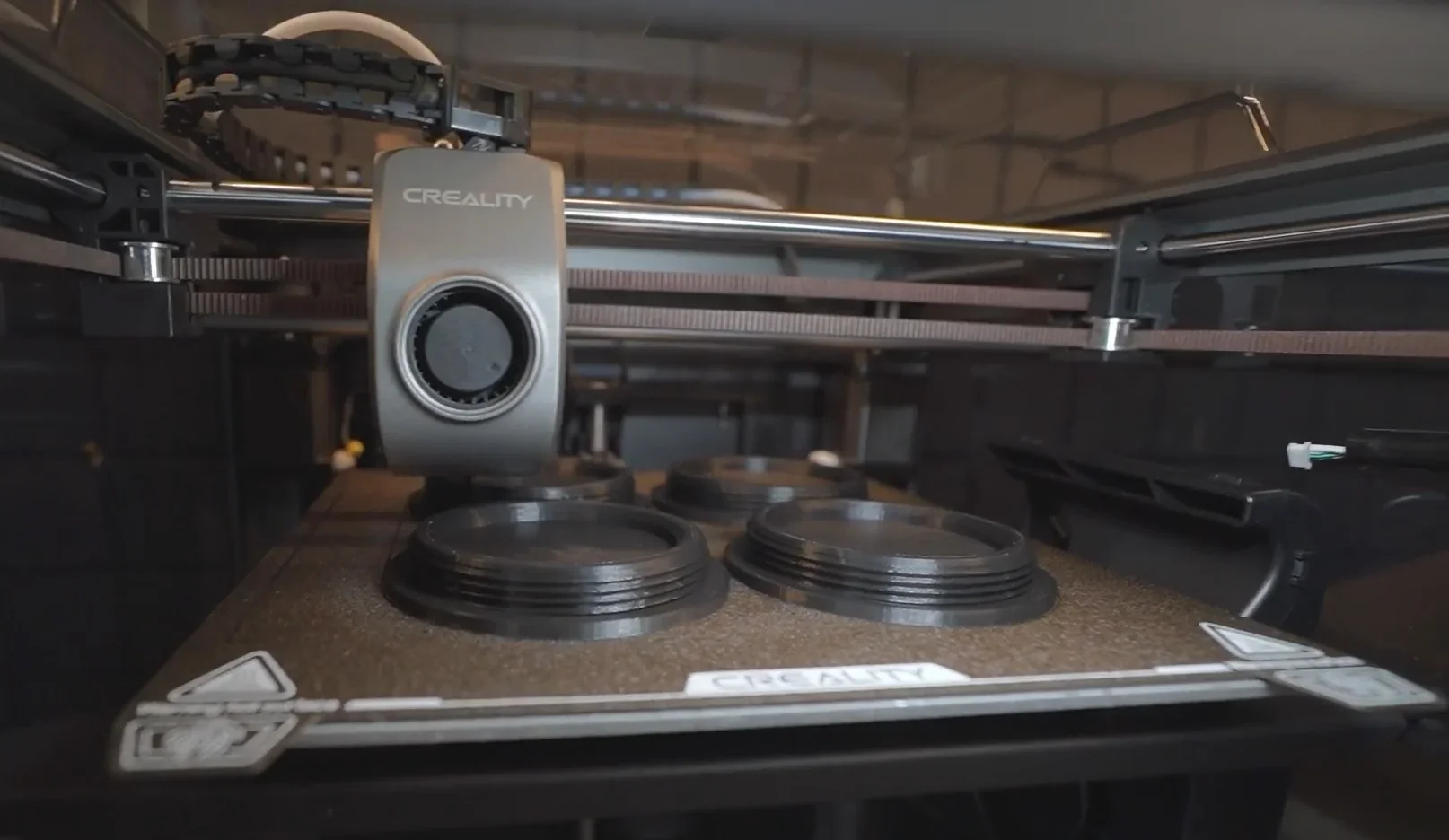 Creality K1 3D printer with extruder head creating circular black parts. Branding visible on plate.