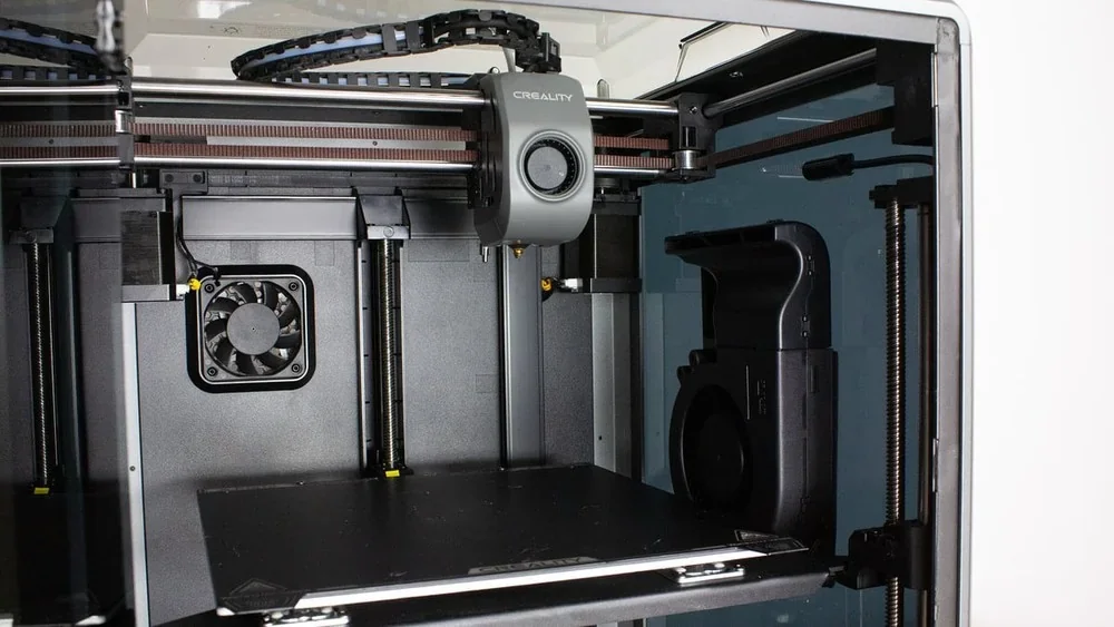 Creality K1 3D printer interior showing extruder, metal frame, cooling fan, and a build plate, highlighting precision engineering and advanced functionality.