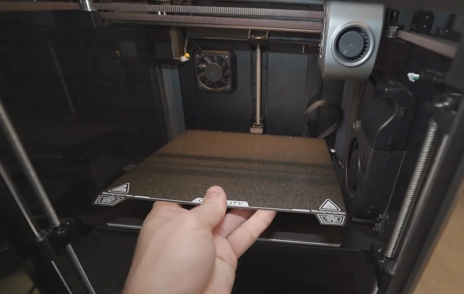 Creality K1 3D printer interior showing a removable build plate being handled, with a focus on the Z-axis and cooling fan inside the printer.