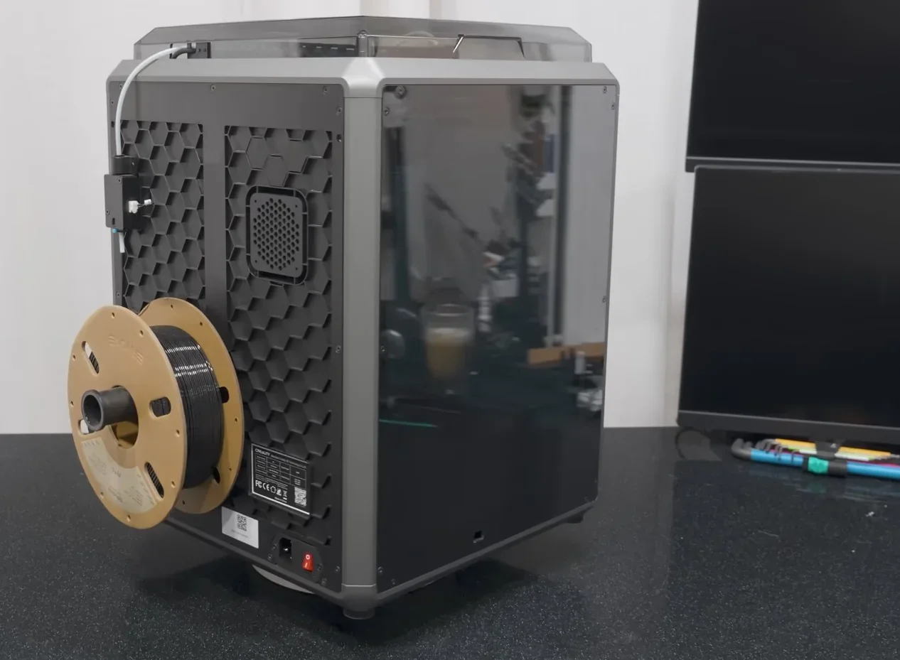 Creality K1 3D printer with a filament spool on the side, featuring a sleek, enclosed design on a workspace, showcasing high-tech capabilities.