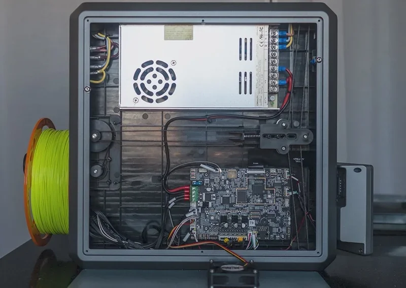 Creality K1 3D printer interior showing circuit board, power supply, wiring, and neon filament reel on the side, illustrating technical components.
