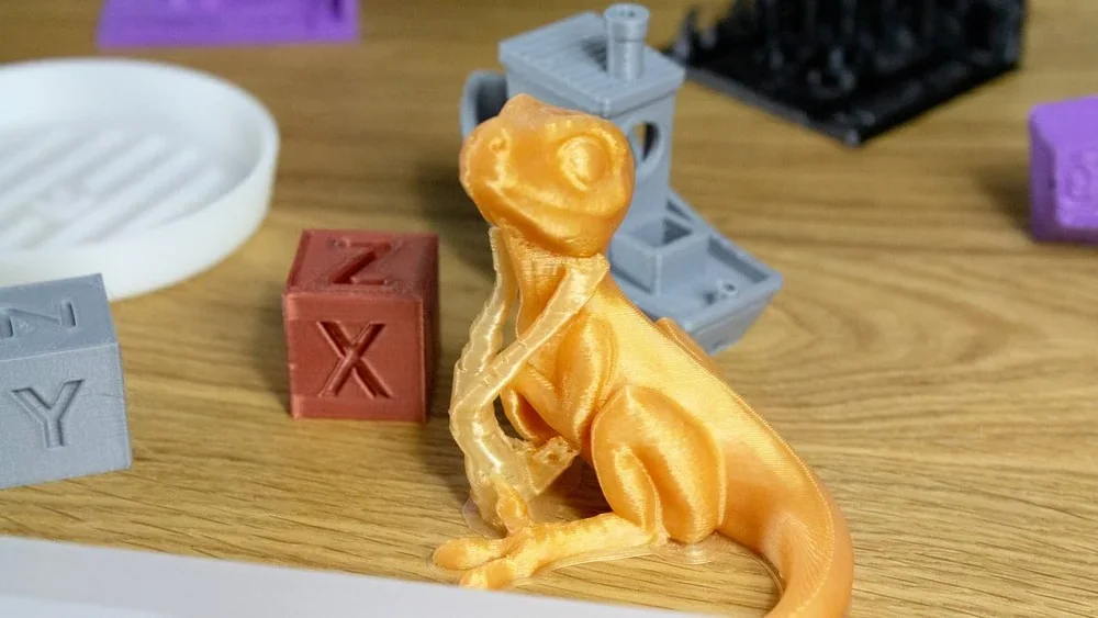 3D-printed orange lizard and colored blocks on a wooden table, created by Creality K1, showcasing detailed print quality and variety of shapes.
