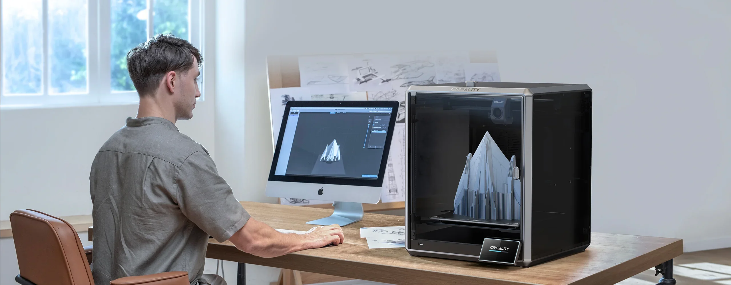 Man designing on computer beside Creality K1 Max 3D printer creating a geometric structure. Bright, modern office with sketching materials nearby.