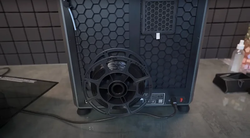 Creality K1 Max 3D printer rear view with honeycomb ventilation, filament spool holder, and power connection on a table in a tiled room.