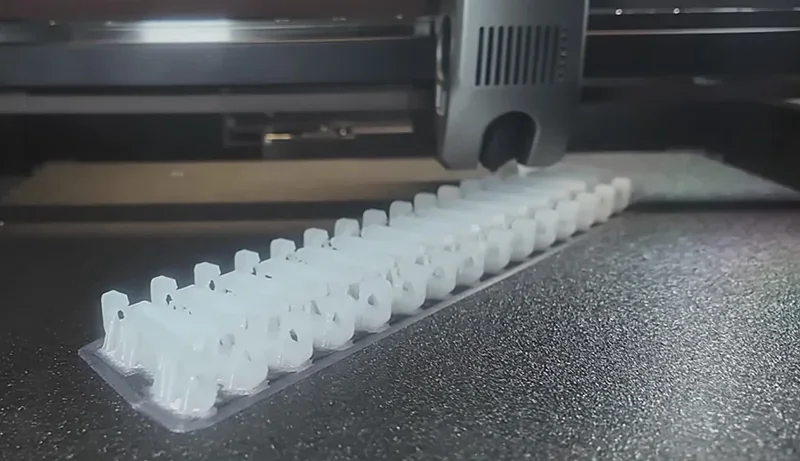 Creality K2 3D printer creating a row of identical white plastic parts on a textured printing bed.