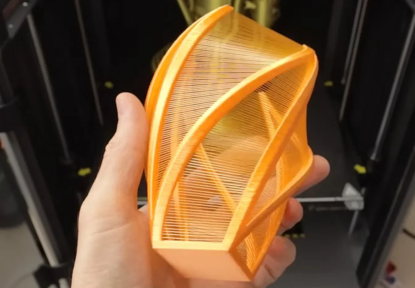 Hand holding an intricate, translucent orange 3D-printed geometrical shape created by Creality K2, showcasing detailed latticework design.