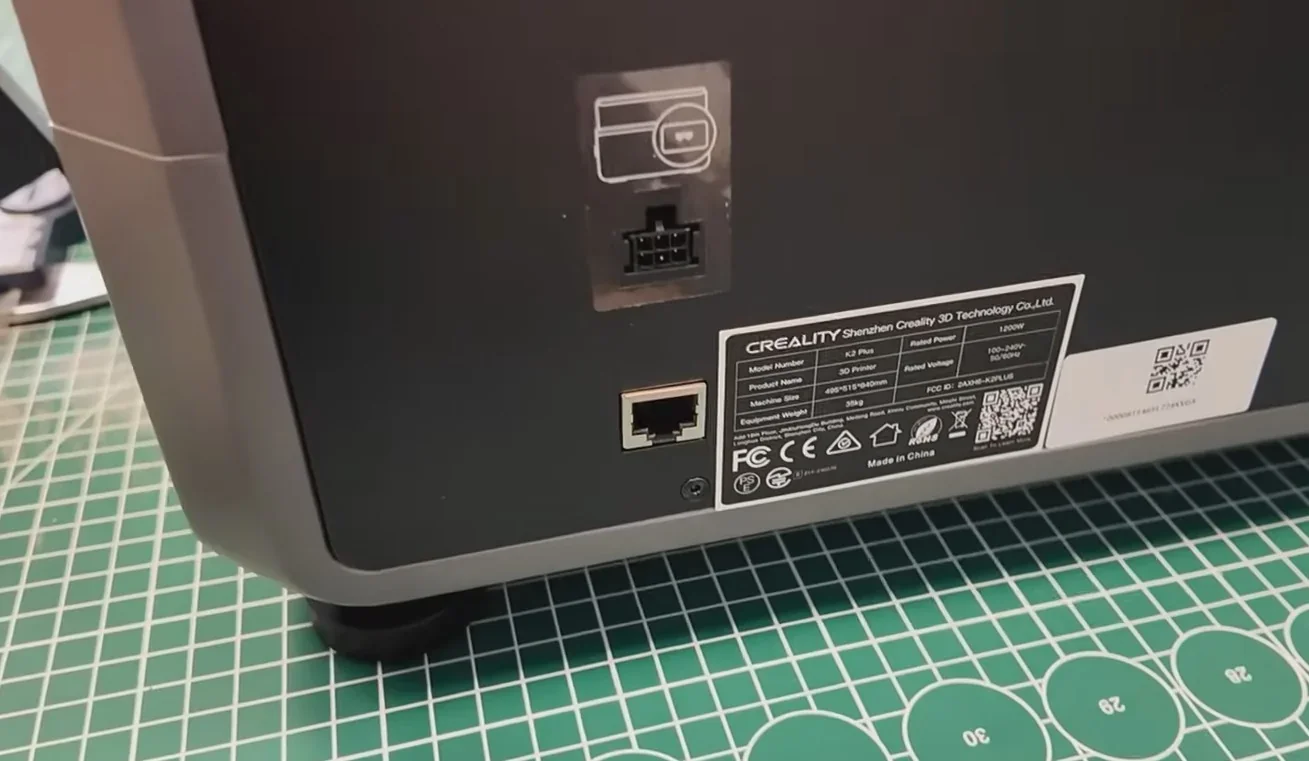 Creality K2 Plus 3D printer back panel showing ports and label on a green grid cutting mat.