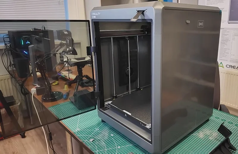 Creality K2 3D printer with open door on a workbench, tools visible in background, showcasing its build volume and design in a workshop setting.