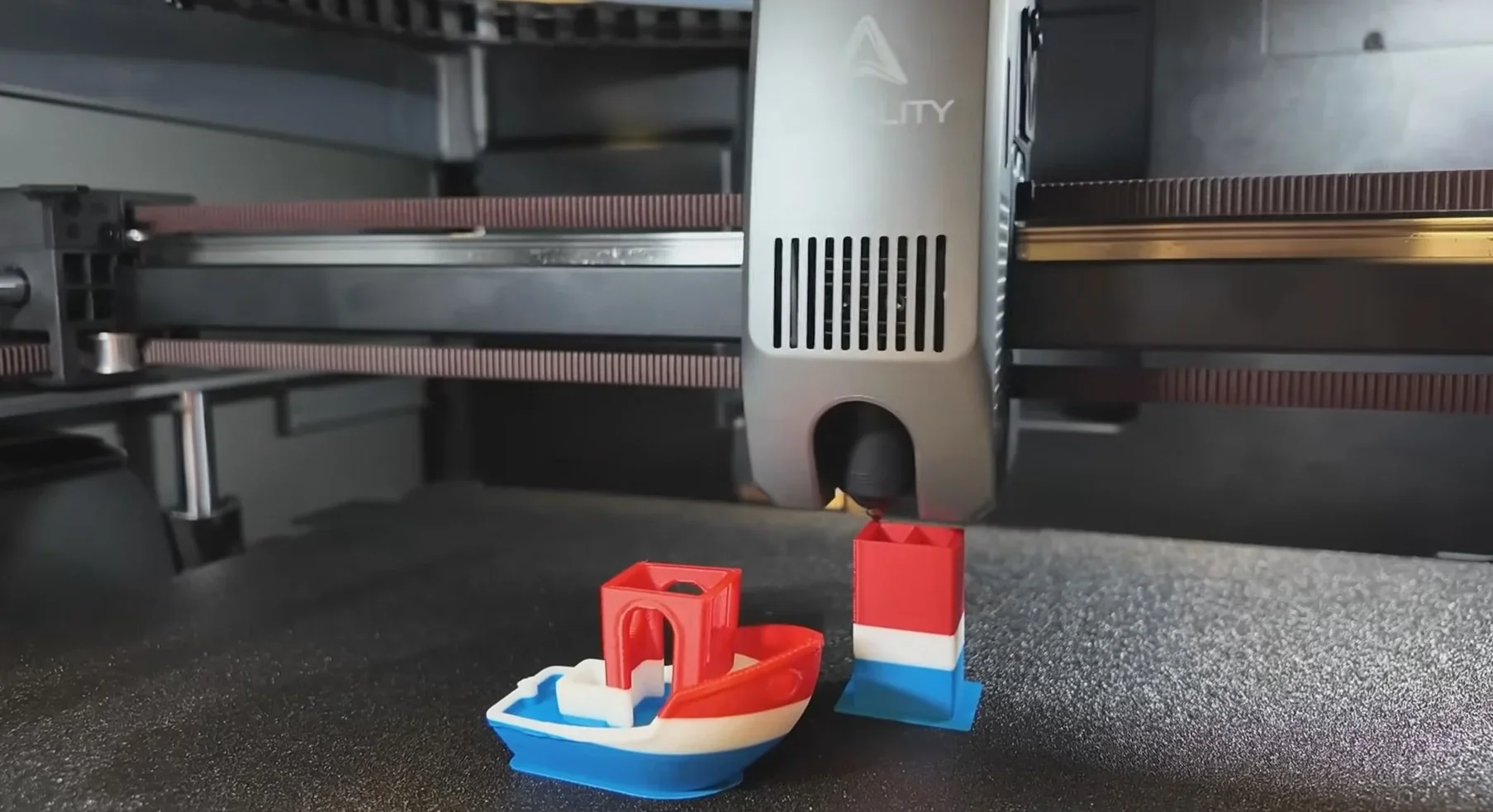 Creality K2 3D printer creating colorful boat and cube objects on textured surface with visible branding and mechanical components.