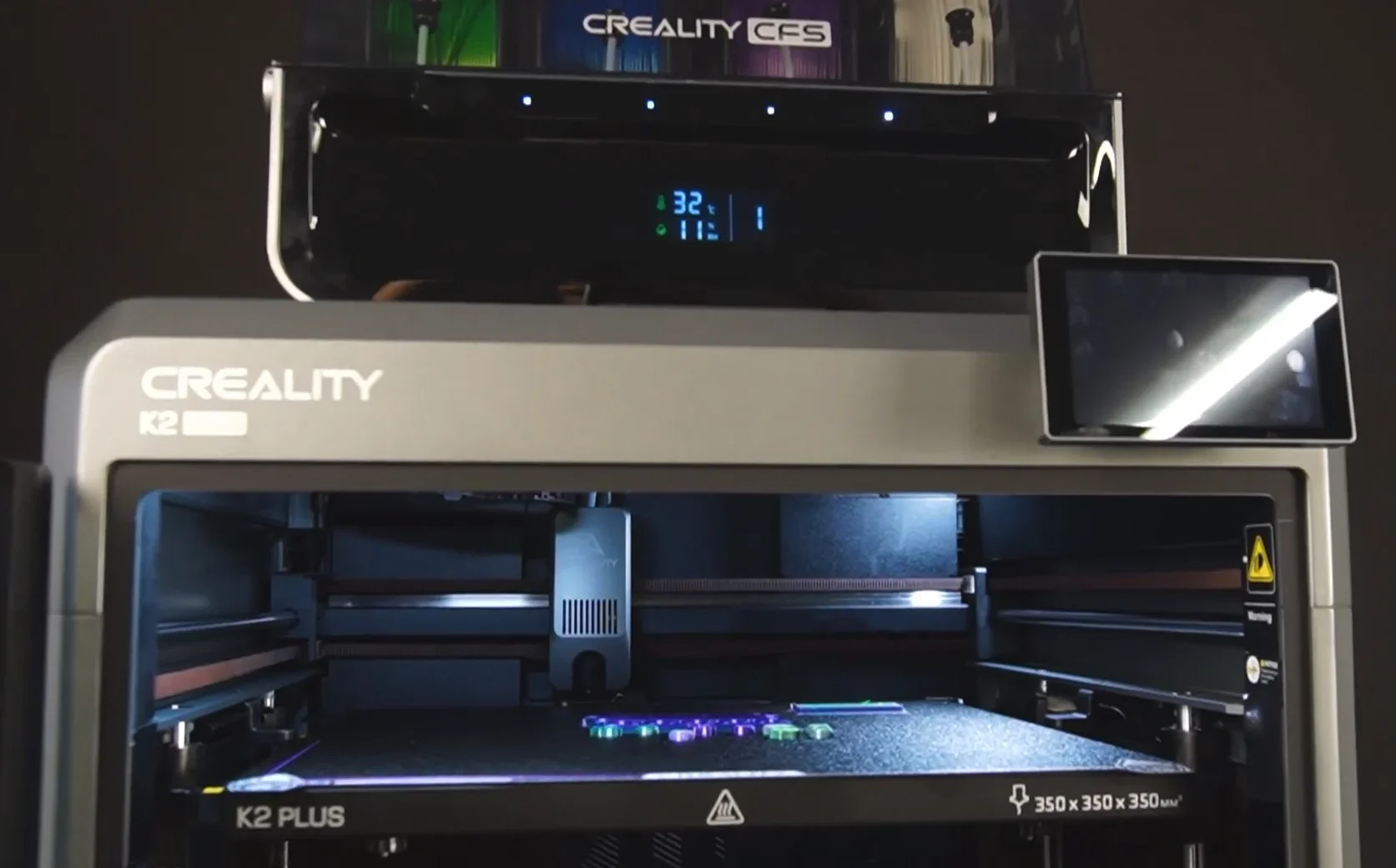 Creality K2 3D printer with display screen, printed parts on the bed, and branding visible; dark background highlights the machine.