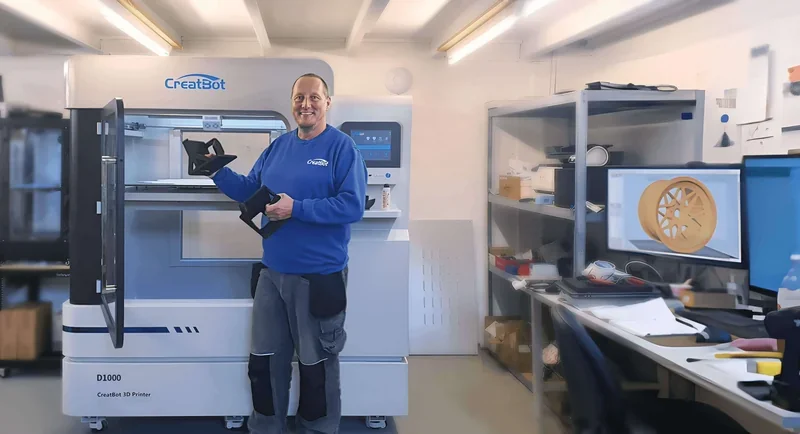 CreatBot D1000 printer in a workshop, man holding 3D printed part. Computer displays 3D model design.