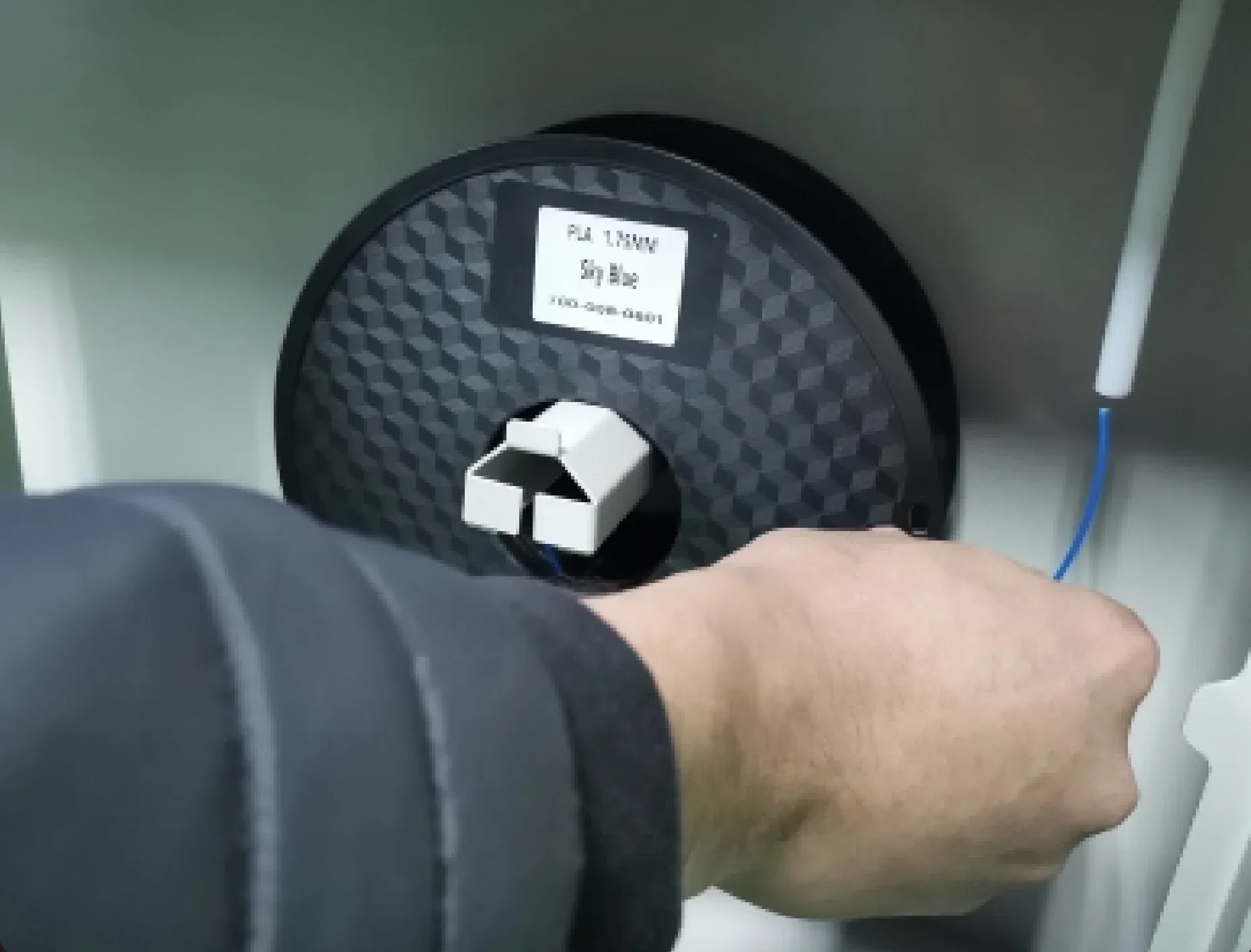 Hand adjusting PLA filament spool on Criatbot D600 Pro 3D printer, showing close-up of geometric texture and label, with blue filament thread.