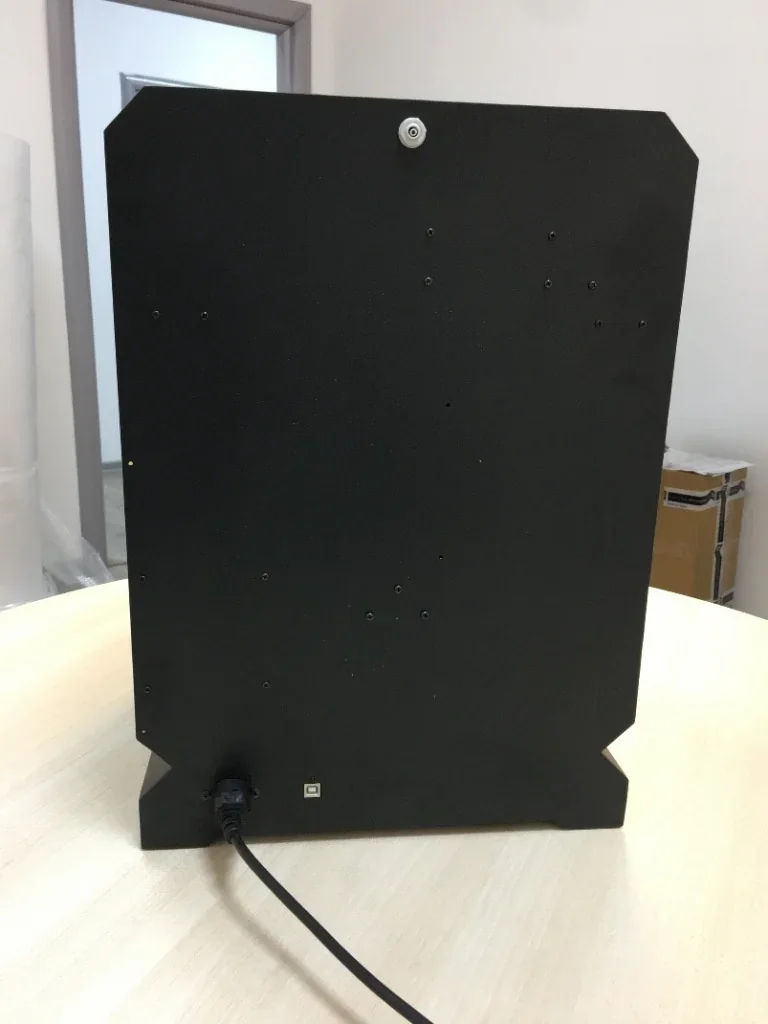 Back view of Creatbot F160 3D printer showing a black panel with a power cable on a light wooden table in a neutral room setting.