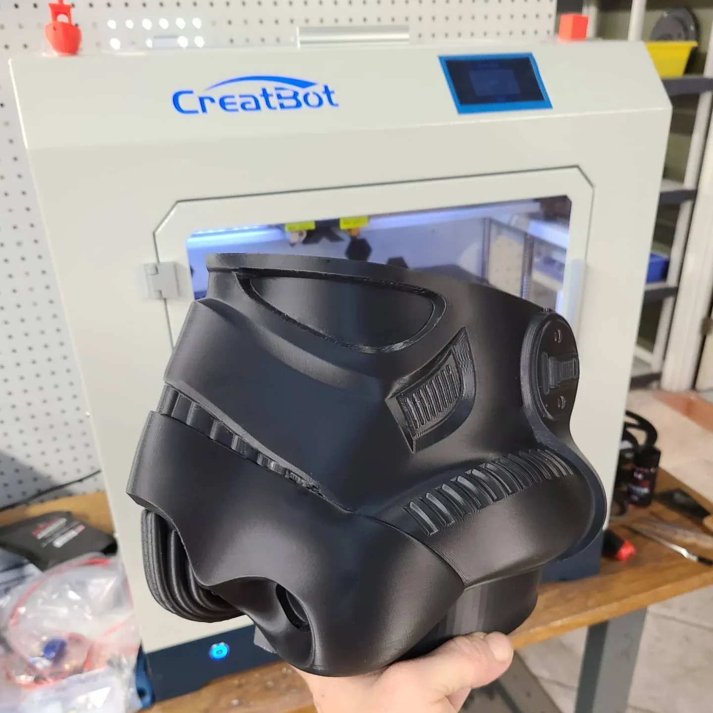 CreatBot F430 3D printer with blue logo, producing a detailed black plastic part, held in hand, on a workspace with tools nearby.