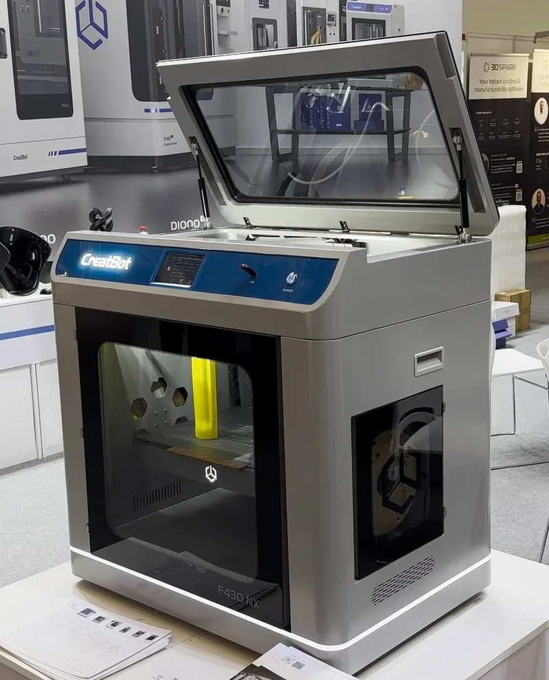 Creatbot F430 NX 3D printer displayed with open lid, showing internal printing area with a yellow part. Branding visible in a trade show setting.