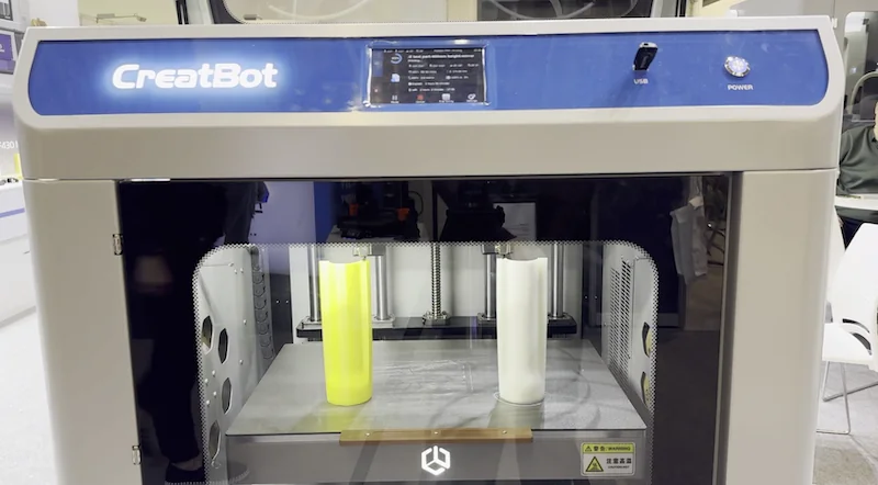 CreatBot F430 NX 3D printer with two printed cylindrical parts, one yellow and one white, showcased inside a sleek, branded enclosure.