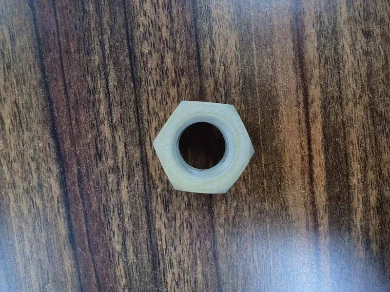 A hexagonal nut sample created by the Creatbot PEEK-250 3D printer. The part is made from a translucent material, likely high-performance plastic such as PEEK, illustrating the printer's capability to produce precise industrial components. The nut is placed on a wood-grain surface, providing contrast and context for the visual detail of the printed object.