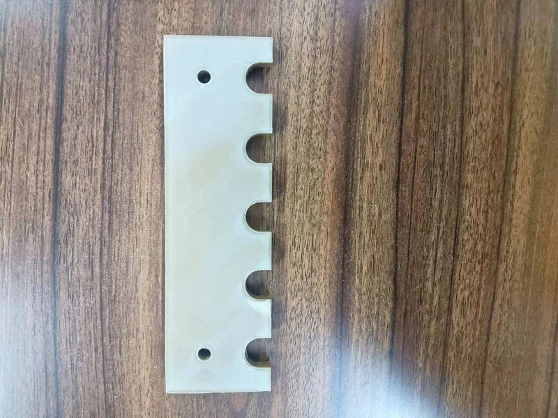 A flat rectangular part created by the Creatbot PEEK-250 sits on a wood-textured surface. The part is designed with six semi-circular indentations evenly spaced along one edge and has two circular holes on opposite corners. The material appears to be a high-grade thermoplastic, emphasizing precision and smooth surface finishes.