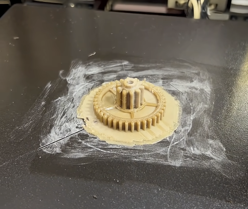 A high-resolution image of a 3D-printed gear part created by the CreatBot PEEK-250. The part is situated on a printing bed, surrounded by a faint layer of residue, possibly from a support or release agent. The gear is finely detailed with visible teeth and a central hub, showcasing the printer’s capability to produce intricate components with high precision.