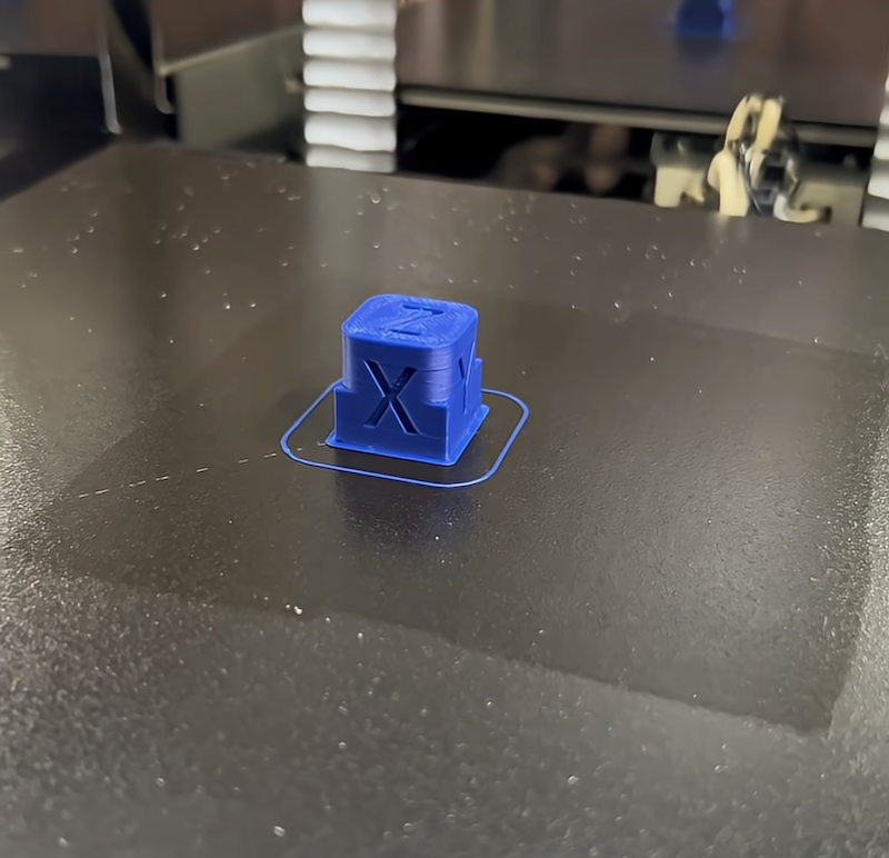 A blue 3D-printed calibration cube with the letter "X" is showcased on a black print bed. This piece was created by the creatbot PEEK-250, demonstrating its precision in producing detailed 3D printed parts. The setting emphasizes the clean surface and printing accuracy of the machine.