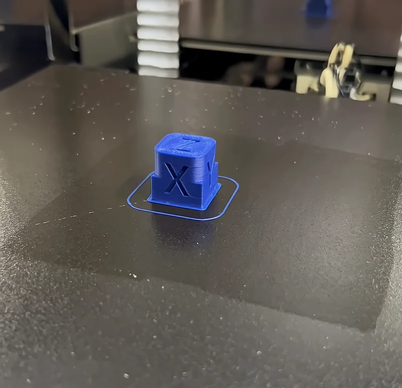 Creatbot PEEK-250 prints a blue cube with an "X" on a black platform, showcasing precision 3D printing capabilities.