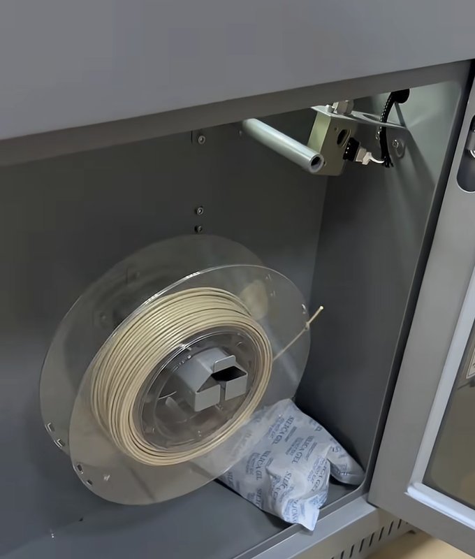 An interior view of a storage compartment within a 3D printer, possibly the creatbot peek-250, showing a spool of filament neatly mounted. The filament is housed on a transparent round spool with a desiccant pouch nearby, likely to maintain optimal printing material conditions. The setting suggests organization and focus on maintaining material quality.