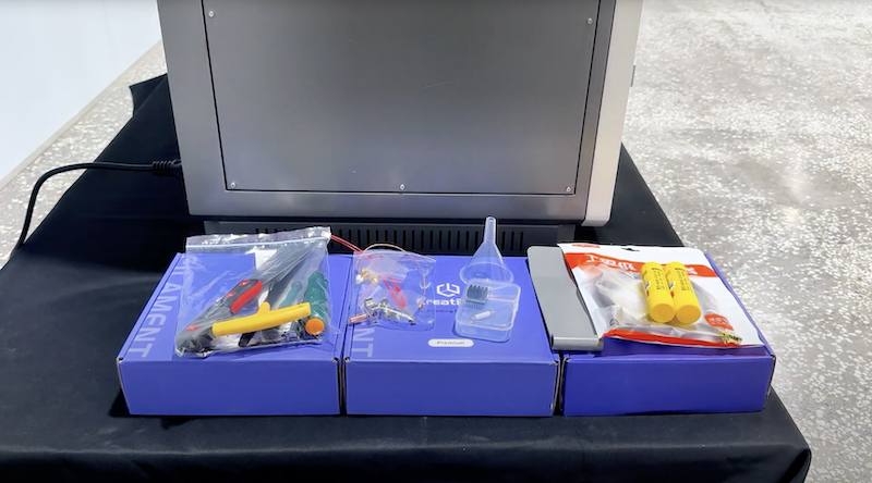 The setup area in front of the CreatBot PEEK-250 3D printer, featuring a variety of accessories and tools on a black tablecloth. Several blue boxes labeled “FILAMENT” are arranged neatly, each containing tools like pliers, a funnel, and silicone lubricant tubes. These accessories are essential for the printer's maintenance and operation, while the printer’s metallic exterior with visible edges provides a sleek industrial appearance.