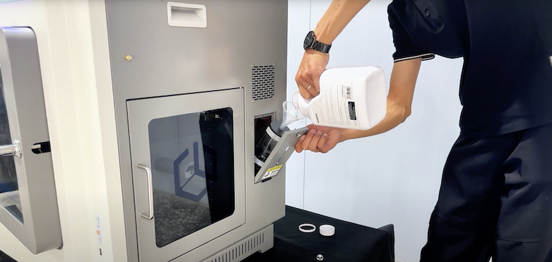 A person is interacting with the creatbot peek-250 printer by pouring liquid into an open compartment on the machine. The printer is partially visible, with a sleek, metallic finish and a clear window displaying the internal components. The individual, dressed in a black outfit, is holding a white container and is equipped with a funnel, suggesting maintenance or the addition of printing materials. The scene implies a professional setting, highlighted by the presence of the creatbot branding on the machine's door.