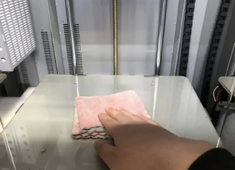 Cleaning glass print bed of Creatbot PEEK-300 3D printer with hand and cloth inside industrial setting.