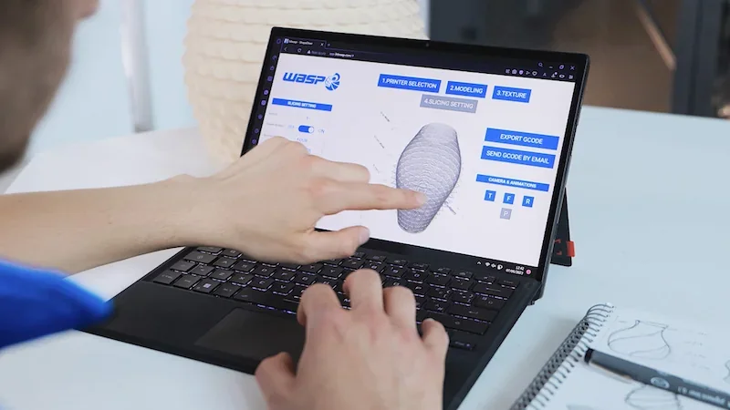 Delta WASP 2040 Clay software on laptop with 3D modeling display; hand interacts with touch screen. Nearby, a 3D-printed clay object and sketch visible.