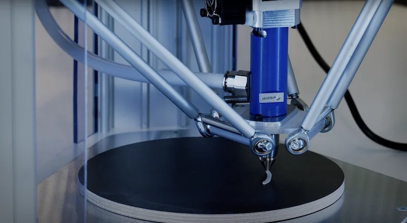 Delta Wasp 2040 Clay 3D printer with intricate nozzle, blue components, metal arms, creating clay print on black circular platform.