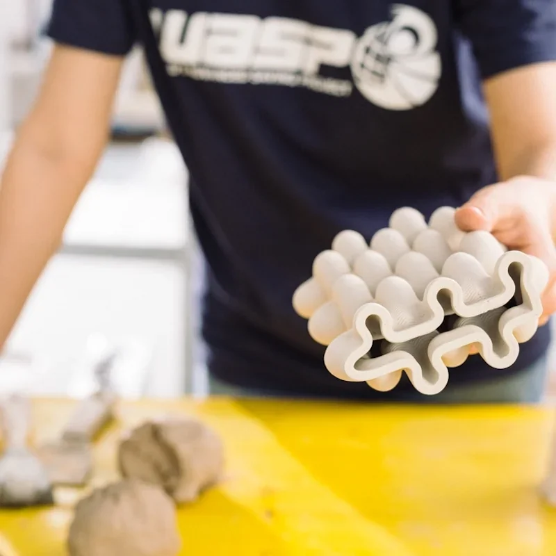 Person holding a clay object with intricate design, created by Delta WASP 2040 Clay 3D printer. Person wearing a Wasp branded shirt in bright workshop.