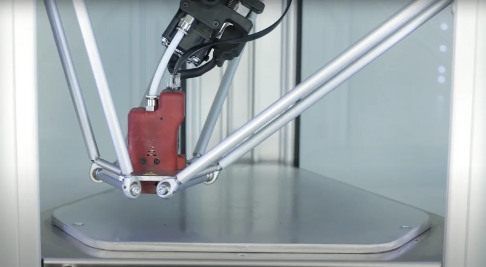 Delta Wasp 2040 Pro 3D printer close-up, showcasing its red extruder with three stabilizing rods, on a metallic print bed inside a white frame.