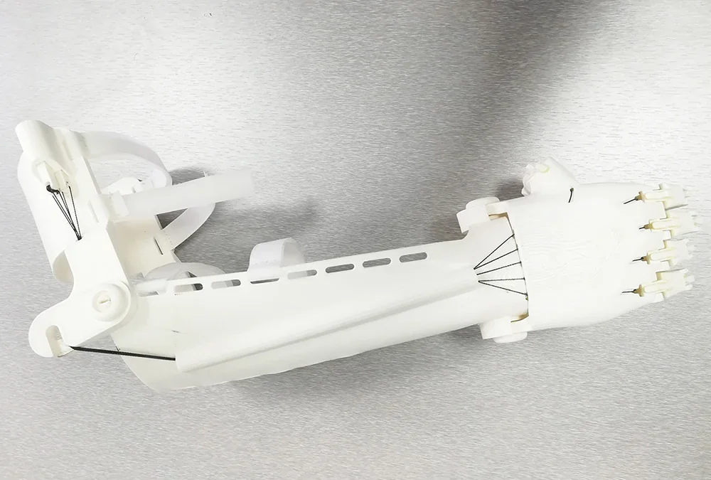 White 3D-printed prosthetic hand, created by Delta WASP 2040 Pro, with articulated fingers, on a metallic surface.