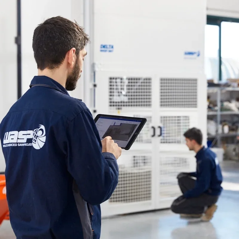 Technicians in a workshop monitor a Delta WASP 3MT 3D printer. One uses a tablet; the printer's branding is visible on its structure nearby.