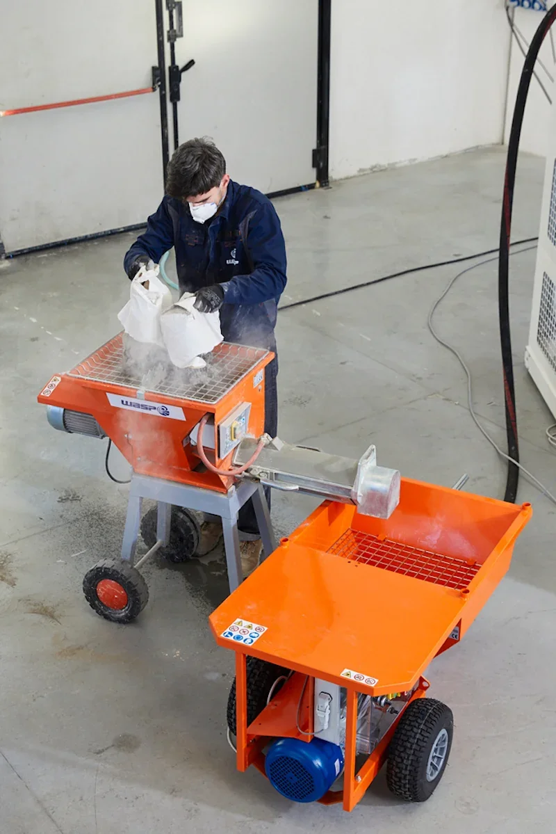 A person in a workshop pours material into an orange Wasp machine for use with the Delta Wasp 3MT 3D printer, featuring visible branding and industrial setting.